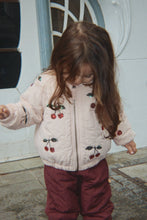 Load image into Gallery viewer, A young child wearing a cream quilted jacket with cherry embroidery and burgundy pants, standing outdoors in front of a white door.
