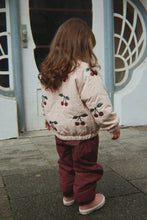 Load image into Gallery viewer, A child viewed from behind wearing a cream quilted jacket with a cherry print and burgundy pants, standing on a paved surface in front of a white building with decorative window frames.