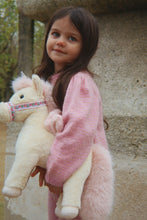 Load image into Gallery viewer, A young girl with long brown hair stands outdoors holding a plush cream-colored stuffed unicorn. She is wearing a fuzzy pink sweater, and the background shows a rustic stone wall.