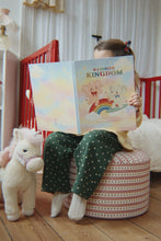 Load image into Gallery viewer, Child reading a book titled 'Rainbow Kingdom' in a cozy room with a crib and stuffed animal.