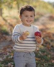 Load image into Gallery viewer, A smiling toddler standing outdoors in a blurred garden or orchard setting, wearing a cream knit sweater with gray horizontal stripes and light gray pants, holding a yellow apple in one hand and a red apple in the other.