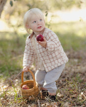 Load image into Gallery viewer, A toddler girl in a beige and white checkered shirt and light gray pants stands outdoors in a grassy, sunlit area, holding a red apple and a small woven basket.