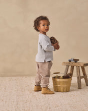 Load image into Gallery viewer, A toddler stands in a rustic studio setting wearing a light gray long-sleeve top, beige pants, and tan boots, holding a pinecone next to a small wooden stool and basket.
