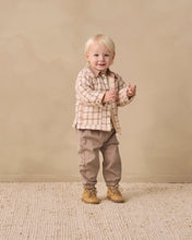 Load image into Gallery viewer, A toddler standing in a studio setting, wearing a beige and white plaid long-sleeve button-up shirt, taupe pants, and tan boots against a warm beige backdrop.