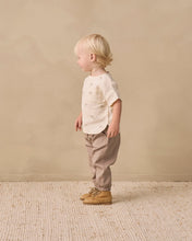 Load image into Gallery viewer, A toddler standing in profile wearing a cream short-sleeve t-shirt with small embroidered motifs, beige pants, and tan lace-up boots, set against a warm beige textured wall and rug.