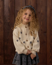 Load image into Gallery viewer, A young girl with curly blonde hair wearing a cream knit cardigan with dark blue embroidered snowflakes and a matching dark blue patterned skirt, standing against a dark wood plank background.