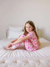 Load image into Gallery viewer, Young girl in pink pajamas sitting on a bed in a bedroom.