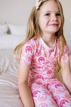 Load image into Gallery viewer, Young girl wearing a pink horseshoe-patterned outfit sitting on a bed.