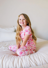 Load image into Gallery viewer, Young girl in pink horseshoe-patterned pajamas sitting on a bed.