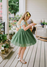 Load image into Gallery viewer, Young girl in a green dress standing on a wooden deck with plants around.