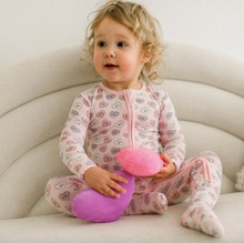 Load image into Gallery viewer, Child wearing a pink onesie with heart patterns, sitting on a white couch holding a purple and pink toy.