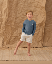 Load image into Gallery viewer, Child wearing a blue shirt and patterned shorts standing against a brown paper backdrop