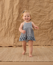 Load image into Gallery viewer, Child in a blue checkered dress standing on a textured brown surface with a brown backdrop