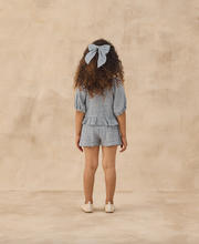 Load image into Gallery viewer, Child wearing a blue checkered outfit with ruffled sleeves and shorts on a beige background