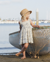 Load image into Gallery viewer, Young girl in a dress and hat standing next to a boat on a beach.