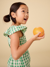 Load image into Gallery viewer, Young girl in a green checkered outfit holding an orange against a beige background