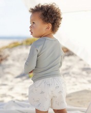 Load image into Gallery viewer, Child wearing a light gray long-sleeve shirt and white shorts with a pattern, standing on a sandy beach.