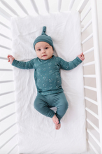 Load image into Gallery viewer, Baby in teal outfit lying in a crib with white bedding