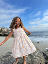 Load image into Gallery viewer, Young girl in a floral dress standing on a rocky beach with ocean and sky in the background