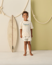 Load image into Gallery viewer, Child wearing a white t-shirt and shorts with a alligator design, standing in front of a surfboard against a light green wall.