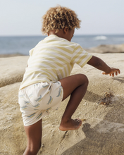 Load image into Gallery viewer, Child climbing on rocks by the ocean wearing a yellow and white striped shirt and patterned shorts.
