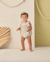 Load image into Gallery viewer, Baby in a floral romper standing against a light-colored wall with decorative elements.