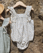 Load image into Gallery viewer, Baby romper with floral pattern on a wooden hanger against a stone wall.