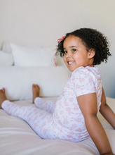 Load image into Gallery viewer, Young girl in a white outfit with pink patterns sitting on a bed.
