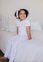 Load image into Gallery viewer, Young girl in a pink floral outfit sitting on a bed.