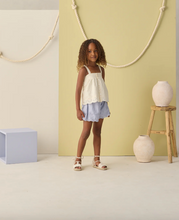 Load image into Gallery viewer, Young girl in a white top and light blue shorts standing in a room with a yellow wall and decorative elements.