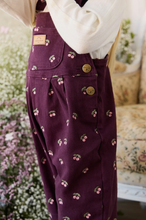 Load image into Gallery viewer, A close-up of a person wearing deep purple corduroy overalls with a cherry print and gold buttons, paired with a cream long-sleeve top in an outdoor garden setting.