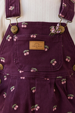 Load image into Gallery viewer, Close-up of a purple denim overall dress with a cherry print and a tan leather patch reading 'JAMIE KAY'.