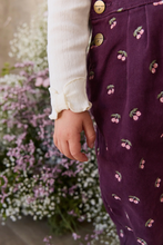 Load image into Gallery viewer, A close-up of a child wearing a dark purple corduroy skirt with a pink cherry print, paired with a cream ribbed long-sleeve top. The background is a soft-focus garden with pink and white flowers.