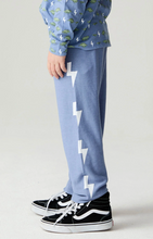 Load image into Gallery viewer, A child wearing light blue sweatpants with large white lightning bolt graphics down the side, paired with a blue dinosaur-print long-sleeve shirt and black high-top sneakers, shown against a light gray studio background.