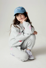 Load image into Gallery viewer, A young girl wearing a light gray sweatshirt and matching sweatpants with small pink and red details, a light blue baseball cap, and gray sneakers, posing in a studio setting.