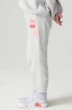 Load image into Gallery viewer, A person wearing light gray sweatpants with pink cherry and bow graphics on the left leg, paired with pale lavender sneakers against a plain light background.