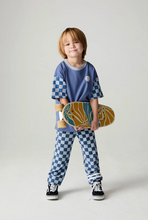 Load image into Gallery viewer, A young child stands against a light gray studio background wearing a blue and white checkered t-shirt and matching checkered pants, holding a skateboard with a yellow and blue pattern.