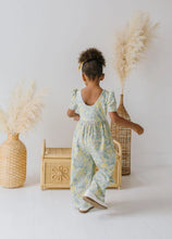 Load image into Gallery viewer, Child wearing a floral dress standing in front of decorative vases and a woven box.