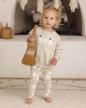 Load image into Gallery viewer, A toddler stands barefoot in a cozy indoor setting, wearing a cream sweatshirt and matching sweatpants with white ghost prints. The sweatshirt features a large white ghost graphic and the text 'Halloween is here.