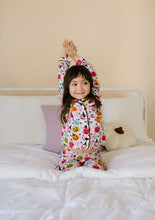 Load image into Gallery viewer, A young girl with dark hair sits on a white bed, stretching her arms overhead. She is wearing a matching long-sleeve crop top and shorts pajama set with a colorful cartoon character print on a white background.