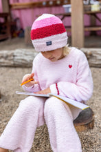 Load image into Gallery viewer, A child wearing a fluffy pink and red striped beanie, a light pink fleece pajama top with a small red heart, and matching pink fleece pants sits on a log outdoors, writing in a notebook with a pencil.