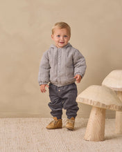 Load image into Gallery viewer, A toddler standing indoors wearing a gray and white gingham quilted puffer jacket, dark gray pants, and tan boots, with mushroom-shaped wooden stools against a beige wall.