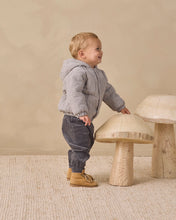 Load image into Gallery viewer, A toddler stands in profile wearing a light gray and white gingham quilted jacket, dark gray pants, and tan boots, standing next to a large wooden mushroom stool in a minimalist studio setting.