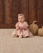 Load image into Gallery viewer, A baby girl wearing a dusty pink dress with white polka dots and a matching pink headband sits on a woven jute rug in front of a dark wood plank wall, with two decorative pumpkins on the right.