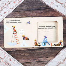 Load image into Gallery viewer, An open children's picture book with illustrated construction-themed pages, photographed from above on a dark wood surface surrounded by a distressed white brick border. The left page shows text about chomping two giant holes, with cartoon workers and tools.