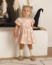 Load image into Gallery viewer, A toddler girl stands indoors wearing a light blush pink dress with white ghost prints, cream knee-high socks, and tan Mary Jane shoes. The background shows a white bench, a black witch hat, and soft seasonal decor.
