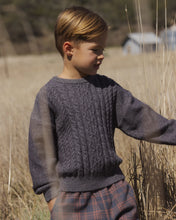 Load image into Gallery viewer, A young boy wearing a dark gray cable-knit sweater and plaid trousers standing in a field of dry grass.

