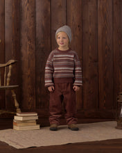 Load image into Gallery viewer, Child wearing a patterned sweater and brown pants standing against a wooden paneled wall.
