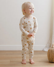 Load image into Gallery viewer, A toddler standing barefoot in a room with light wood floors and a cream wall, wearing a cream long-sleeve pajama set with a tan pumpkin print.
