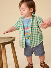 Load image into Gallery viewer, Child wearing a green checkered shirt over a colorful graphic t-shirt, standing next to a wooden stool.
